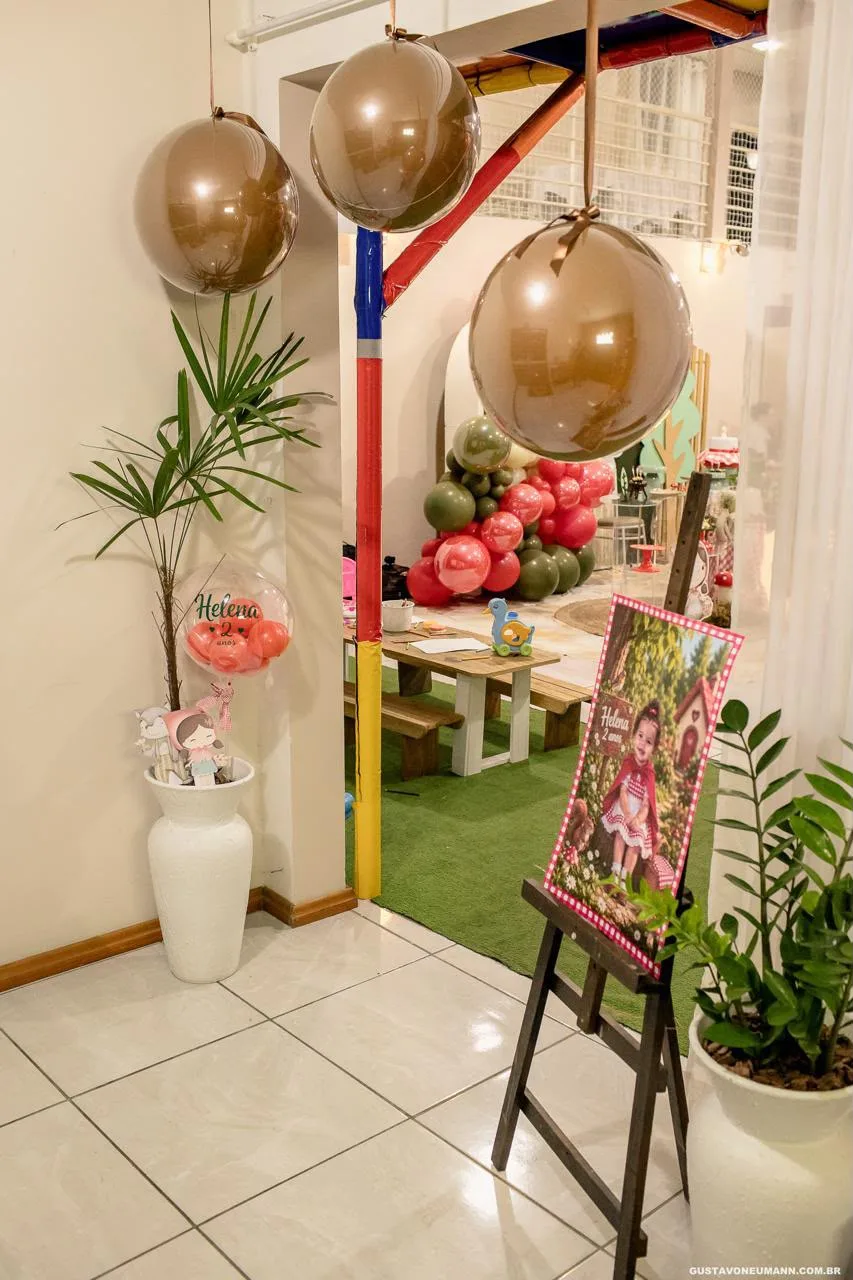 Entrada do salão decorada com balões pendurados no teto, vaso com balão de aniversário, cavalete com painel e mesa de madeira sobre grama sintética