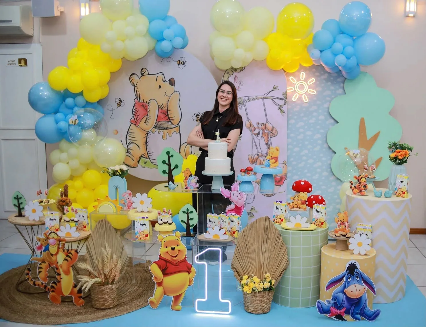 Mesa de festa no tema do Ursinho Pooh montada no Madê Kids, com painel ilustrado, arco de balões e o número 1 em neon, e uma pessoa em pé ao lado da mesa