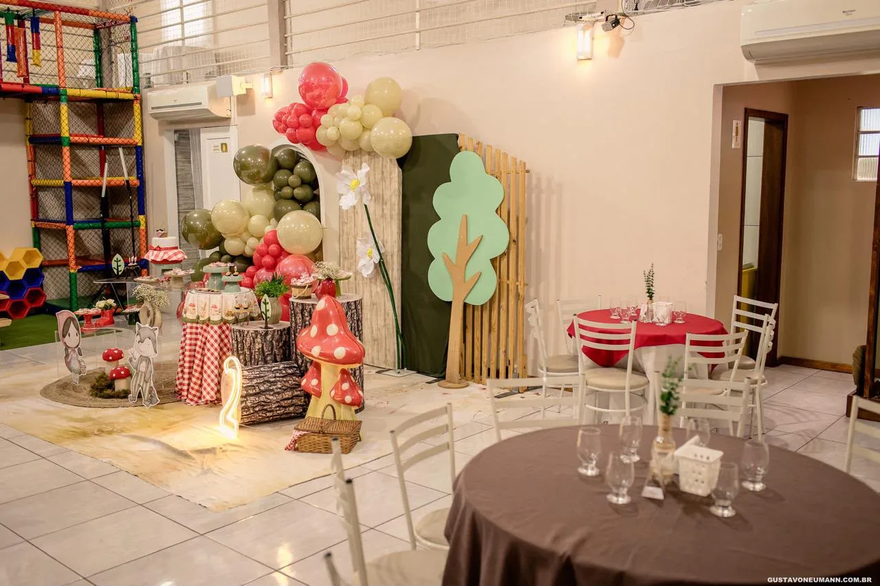 Salão do Madê Kids montado para uma festa com tema de floresta: painel em arco com árvore e cogumelos, arcos de balões em vermelho e verde, mesa decorada e o brinquedão ao fundo